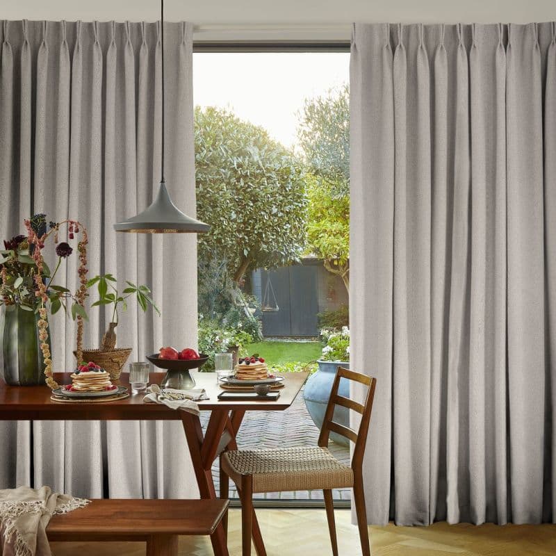 Pleated, floor-length grey curtains drawn partly open to frame a central glass door, filtering soft daylight. Dining table with breakfast plates and pendant lamp overlooks a leafy backyard garden.