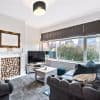 Dark gray Roman blind lowered across a wide front window, partially filtering daylight; living room with tufted gray sofa, TV, coffee table and decorative stacked logs in the fireplace, hedge and houses outside visible.
FOR SALE