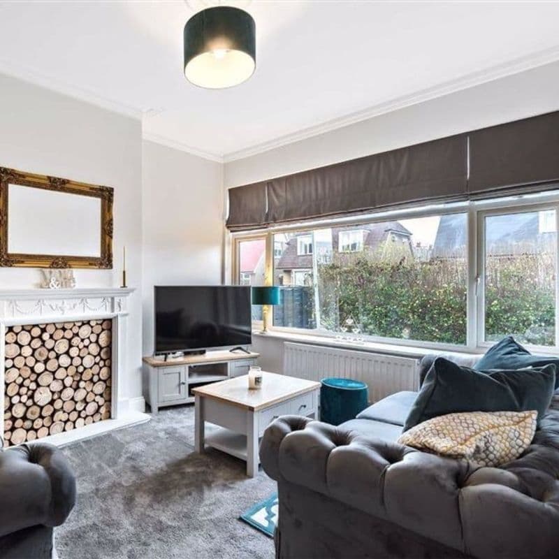 Dark gray Roman blind lowered across a wide front window, partially filtering daylight; living room with tufted gray sofa, TV, coffee table and decorative stacked logs in the fireplace, hedge and houses outside visible.
FOR SALE