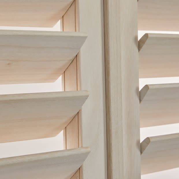 Light unfinished wood plantation shutters with angled horizontal louvers and a central stile — louvers tilted open, filtering soft daylight — close-up interior view of adjacent panels.