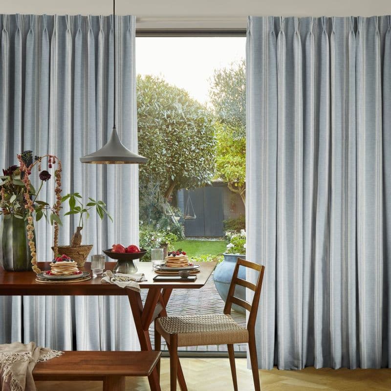 Floor-to-ceiling pleated light-gray curtains, parted to reveal a glass patio door; curtains soften incoming daylight. Wooden dining table set with breakfast plates and pendant lamp, opening onto a sunlit garden.