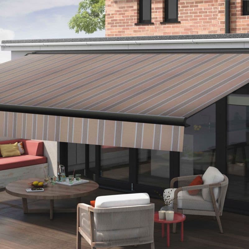Striped beige retractable awning with matching front valance, extended and shading a patio — outdoor seating on a wooden deck before glass doors and a brick upper wall, daylight.