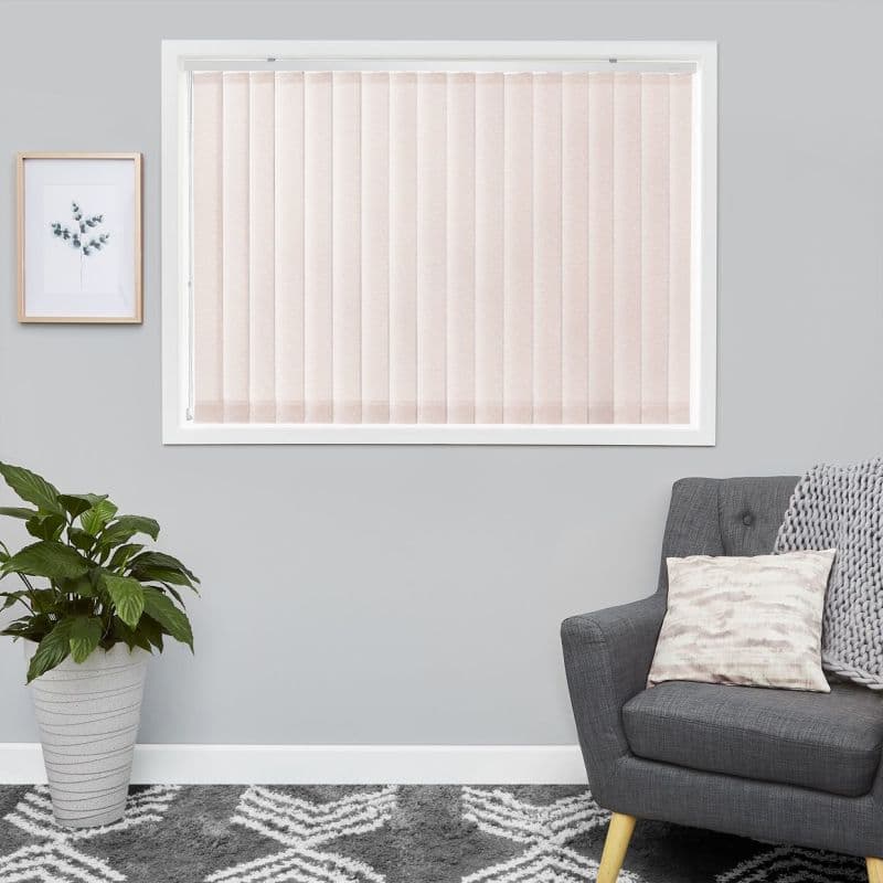 Pale pink vertical blinds covering a white-framed window, closed and softly filtering light; situated on a light-gray living-room wall above a patterned rug, beside a potted plant and gray armchair.