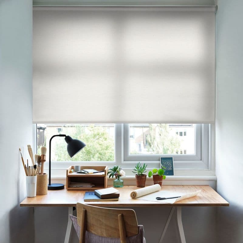 A white fabric roller blind, lowered across a double window, lightly diffusing sunlight over a tidy wooden desk with a lamp, art supplies, small potted plants and a suburban view.