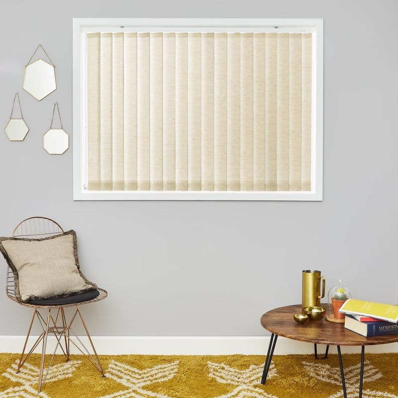 Beige vertical blinds, fully closed, softly filtering light; in a white-framed window above a living-room corner with gray walls, wire chair with cushion, round wooden table and mustard rug.