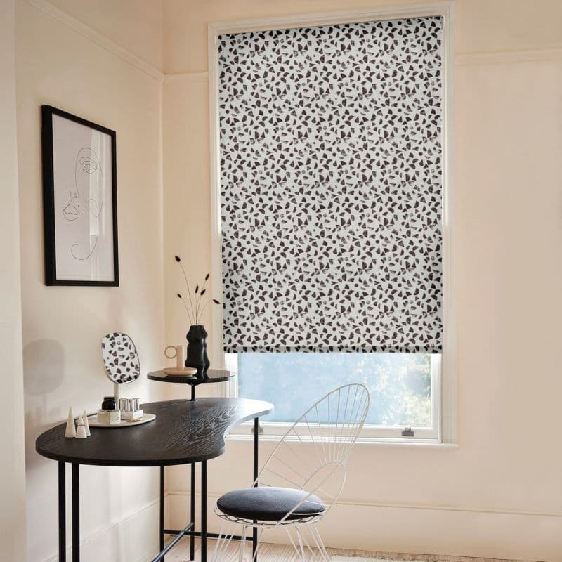 Patterned roller blind (white with scattered black terrazzo-like shapes) fully lowered, filtering daylight into a pale, minimalist room with a small black desk, wire chair, and framed line-art.