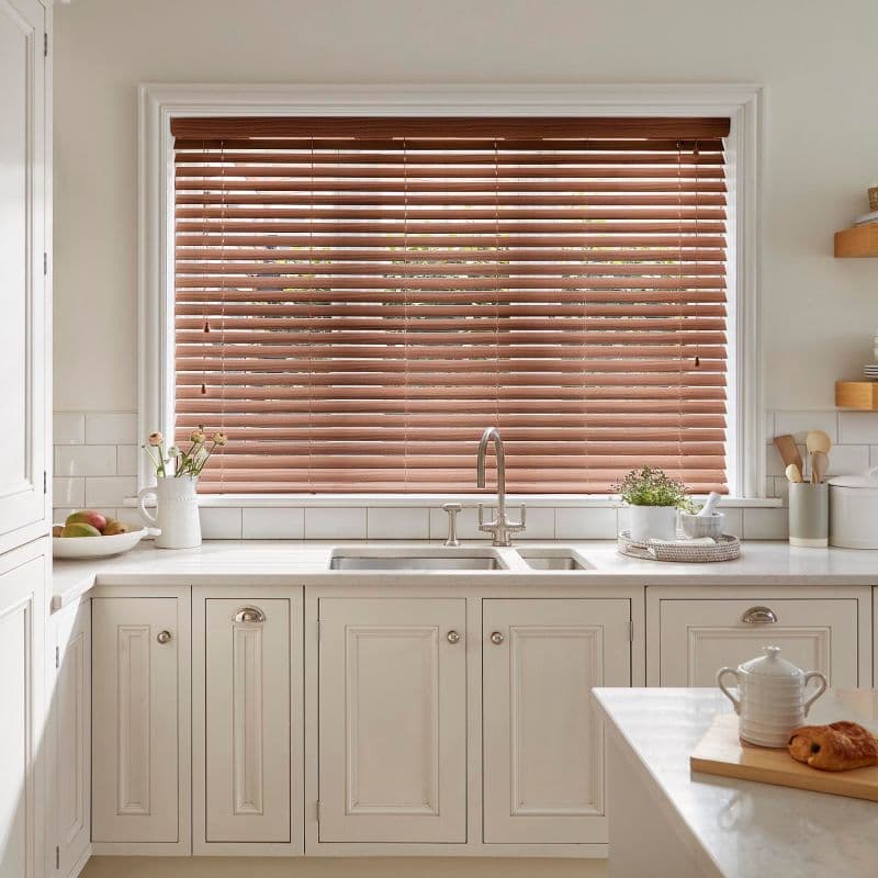 Wooden horizontal blinds covering a large central window, slatted and lowered, slightly tilted to filter daylight; set above a farmhouse sink on a cream kitchen countertop with plants and utensils.