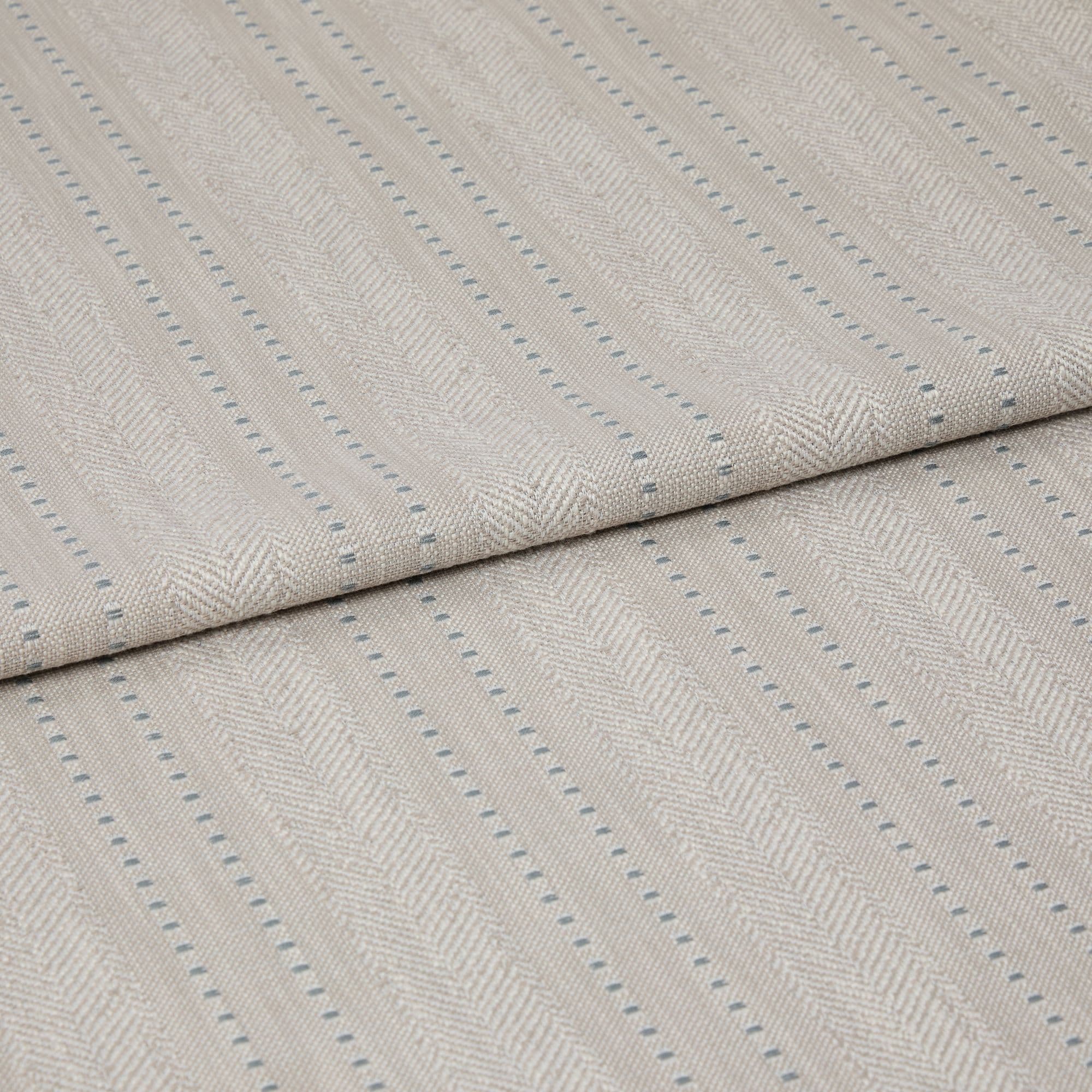 Beige woven fabric featuring herringbone chevrons and thin blue dotted vertical stripes — folded over itself, displayed as a close-up textile sample on a neutral, softly lit surface.