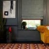 A gray Roman shade with red trim, partially lowered and filtering daylight through a sash window; in a dark-gray living room beside a mustard sofa, cactus and abstract painting.