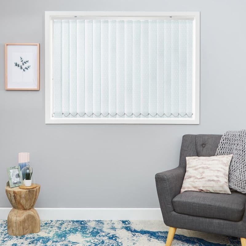 White patterned vertical blinds cover a rectangular window, fully closed with scalloped hem, filtering soft daylight in a gray-walled room with gray armchair, knit throw, side table and blue rug.