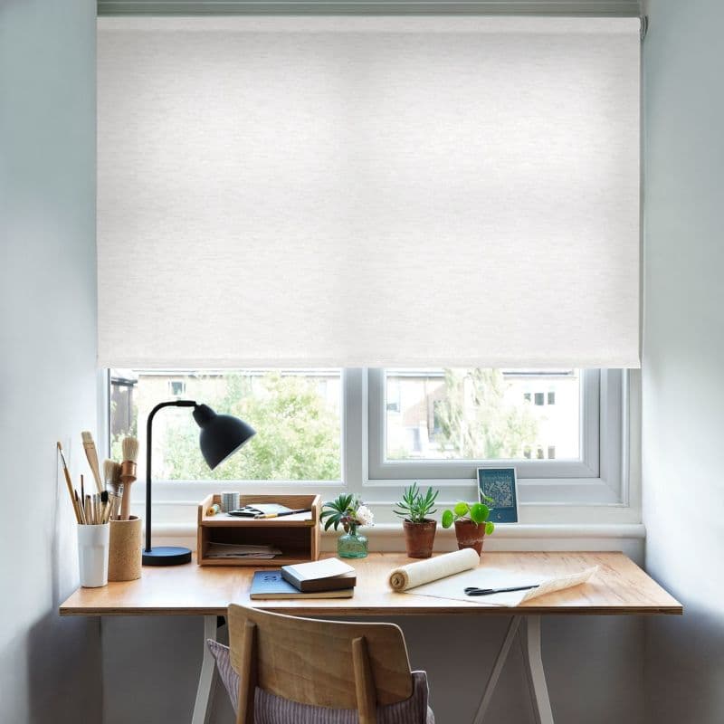 White fabric roller blind lowered over the window, diffusing daylight; above a wooden desk with a black task lamp, art brushes, notebooks and small potted plants in a bright workspace.