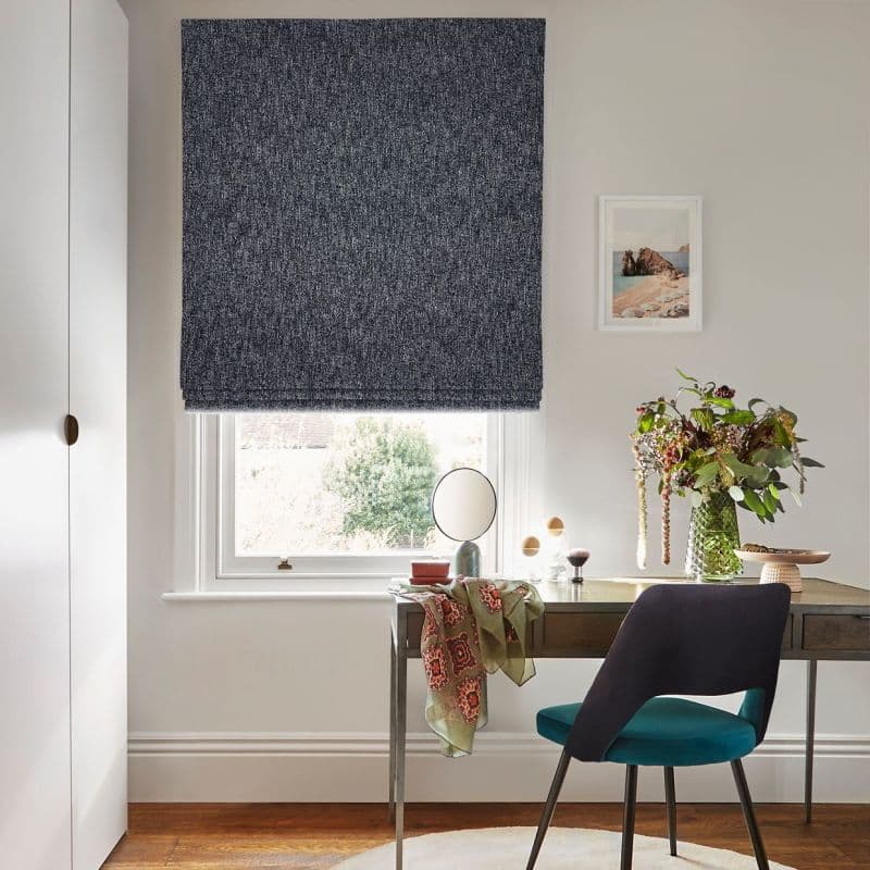 Textured navy Roman blind, lowered fully, filtering light through a narrow bottom gap; set over an office window above a desk with chair, mirror, vase of flowers and framed print.