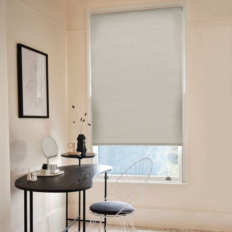 Light-gray textured roller blind, fully lowered with a narrow bottom gap, filtering soft daylight; set in a pale-walled minimalist home office with black curved desk, wire chair, and framed line-art.
