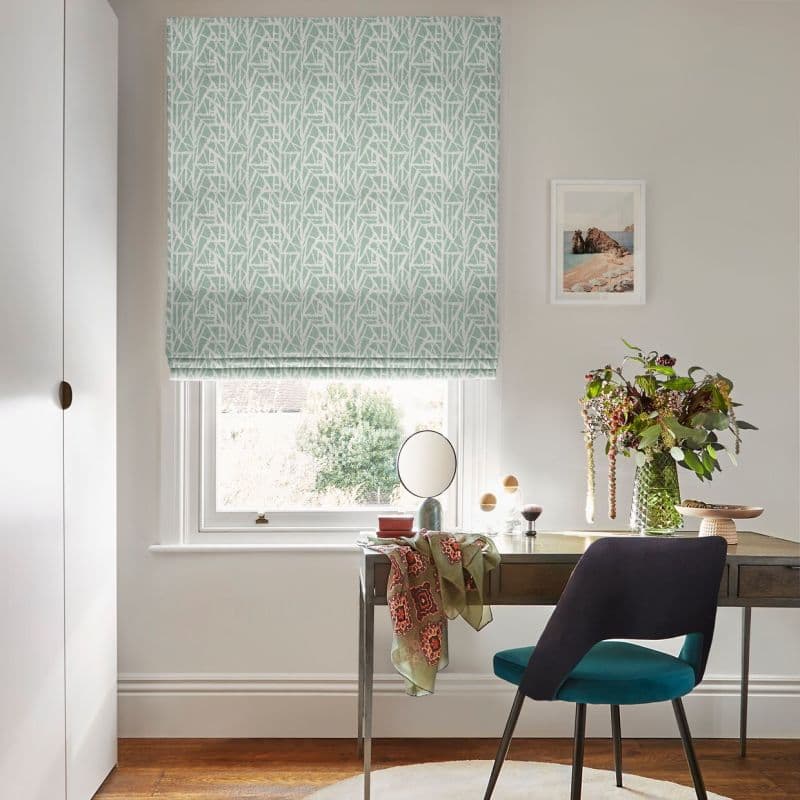 A patterned mint-green Roman blind covers the window, lowered and gently filtering daylight; a wooden desk with mirror, vase of flowers and teal-upholstered chair in a bright, neutral home office.