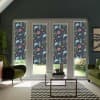 Floral-patterned roller blinds cover tall French doors and sidelights, blocking light in a modern living room with green armchairs, glass coffee table, chevron rug and glimpses of a garden.