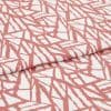 White fabric with coral-pink angular, brushlike linear pattern, folded over itself; fabric lies flat, close-up studio view showing texture and repeating print.