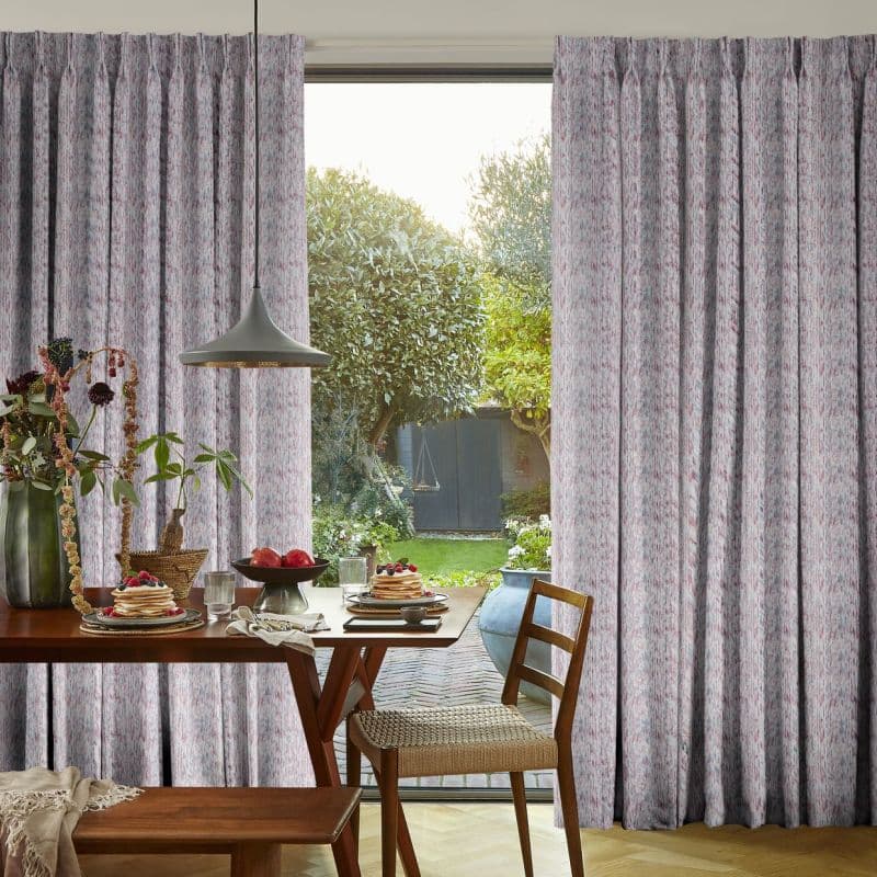 Floor-length pleated patterned curtains, pale grey with pink flecks, partially parted to frame a sliding glass door, filtering soft daylight over a breakfast table and lush garden view.