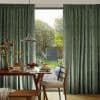 Floor-length, pleated green curtains hung on a ceiling track, partially drawn to frame a glass door; sunlit backyard visible beyond, dining table set with pancakes and wooden chairs inside.