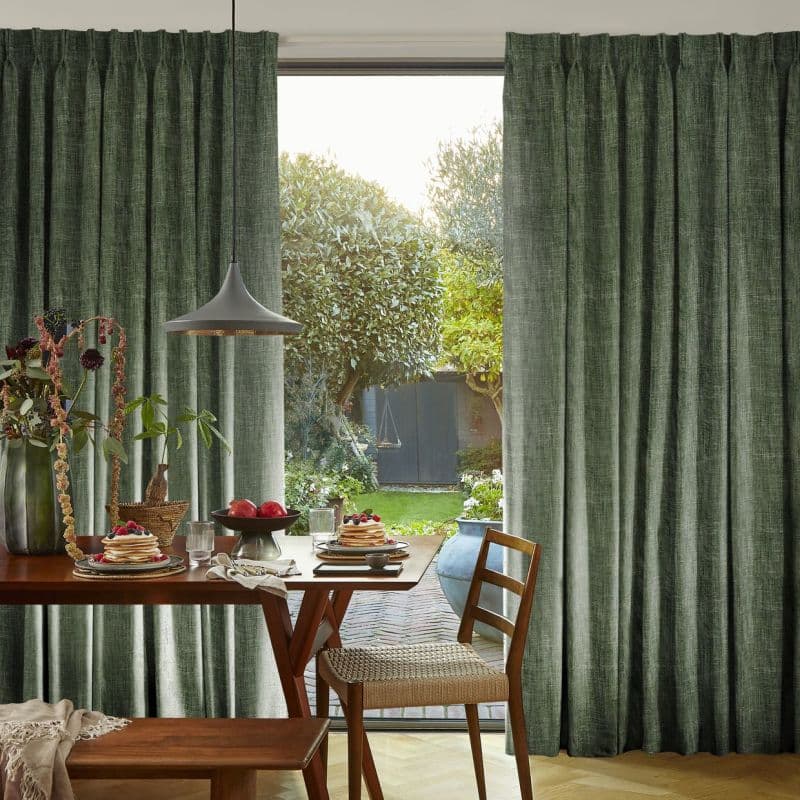 Floor-length, pleated green curtains hung on a ceiling track, partially drawn to frame a glass door; sunlit backyard visible beyond, dining table set with pancakes and wooden chairs inside.