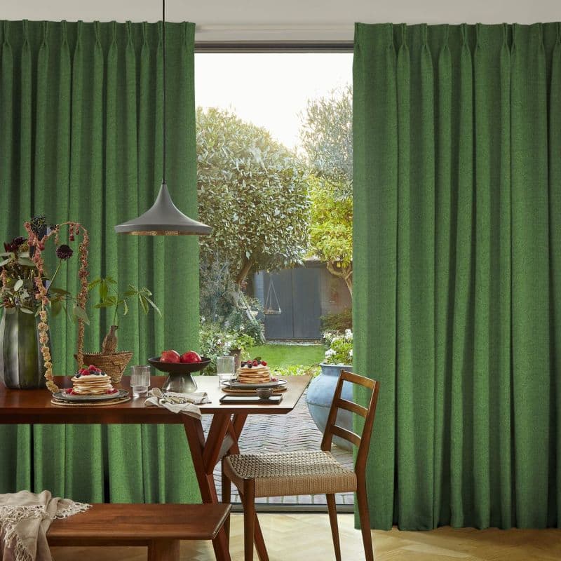 Green pleated floor-length curtains pulled aside centrally, textured fabric framing a sunlit backyard with trees and shed; a dining table with pancakes and a pendant lamp fills the foreground.