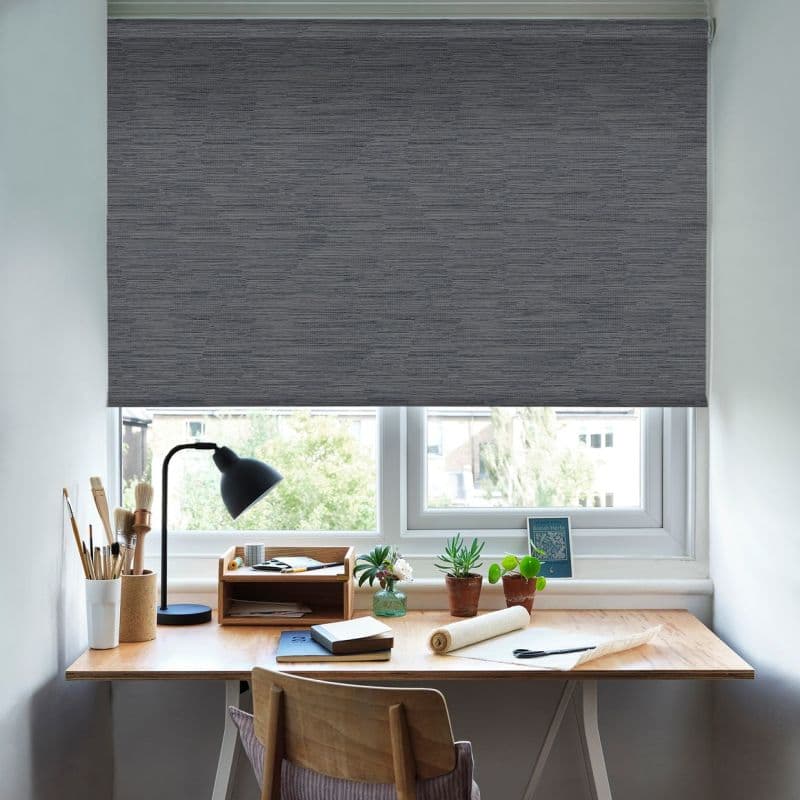 Gray textured roller blind, lowered to cover the window, filtering daylight. Home desk workspace beneath with lamp, potted plants, art supplies and chair; outside view of trees and buildings.