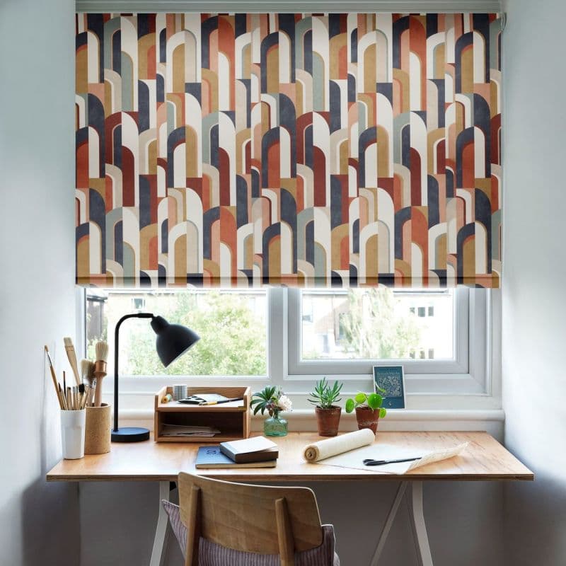 A multicolored roller blind with overlapping arch motifs, lowered across a double window, filtering daylight. Below, a wooden desk holds a lamp, brushes, notebooks and potted succulents, bright workspace.