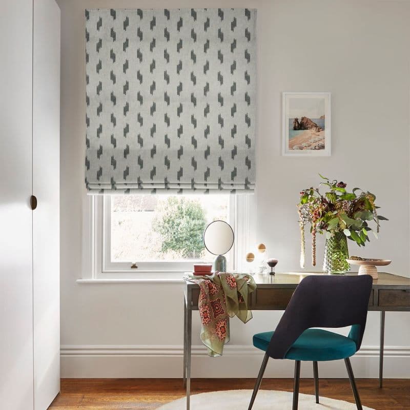 Patterned grey Roman blind with small dark geometric motifs, lowered to just above the sill, filtering soft daylight over a compact home study: wooden desk, teal chair, mirror and vase of flowers.