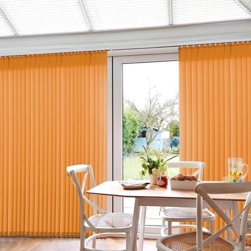 Orange vertical blinds mostly closed, parted slightly at center over a sliding glass door, in a dining nook with white chairs and table, garden visible outside.