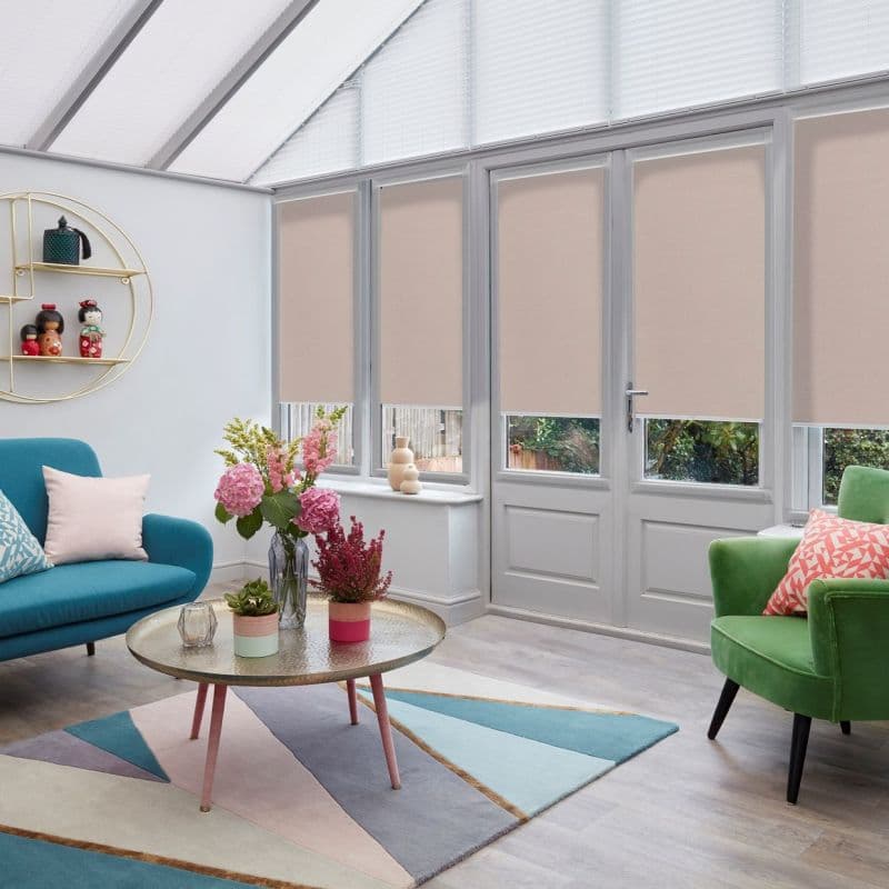 Beige roller blinds lowered over tall glass doors and triangular skylights, partially blocking and diffusing daylight; inside a bright conservatory-style living room with colorful sofas, coffee table, and floral arrangement.