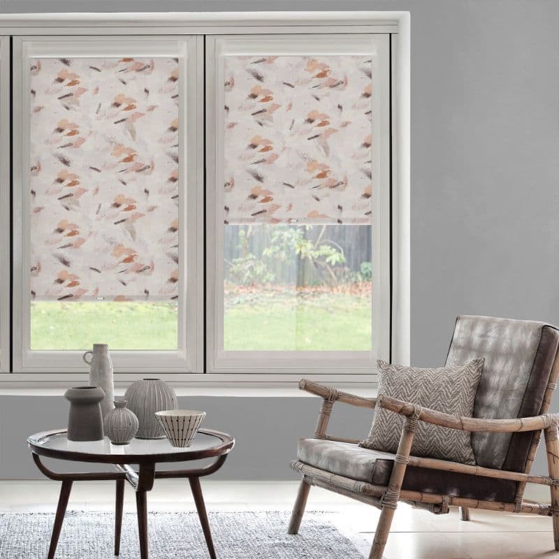 Patterned roller blinds with soft pink-beige brushstroke design cover two adjacent windows—left fully lowered, right halfway—filtering daylight and partially revealing grassy backyard; living room with chair and coffee table.