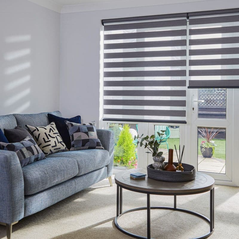 Dual-layer roller blinds with alternating opaque and sheer horizontal stripes, lowered over patio doors and windows, filtering sunlight; living room with blue sofa, patterned cushions, coffee table, garden view.