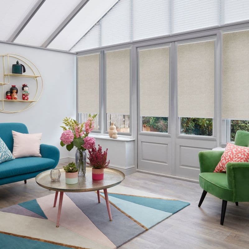 Beige roller blinds on tall French doors and windows (pleated roof blinds above), partially lowered and filtering daylight in a bright conservatory with pastel sofas, coffee table and garden view.