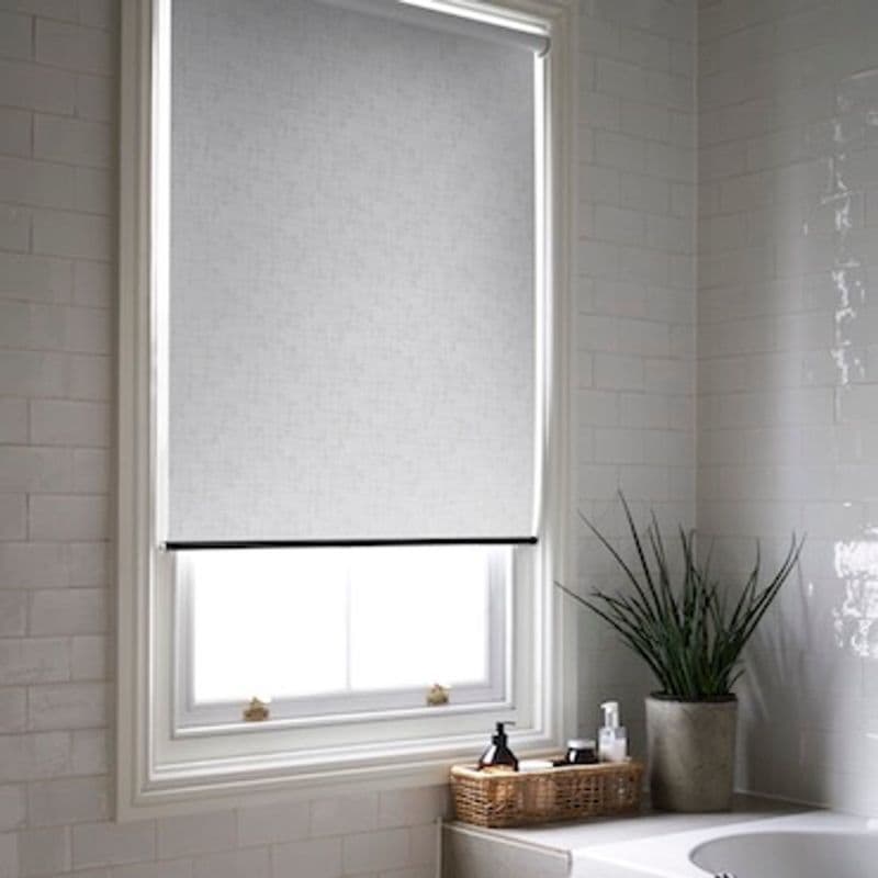 Grey fabric roller blind, lowered and covering most of the window with slight light seep at bottom; filtering daylight in a tiled bathroom above bathtub, with a potted plant and basket.
