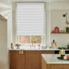 A white textured roller blind, fully lowered across a sash window, diffusing daylight over a kitchen sink with brass faucet, marble countertop, wooden cabinets, open shelf and brick courtyard view.