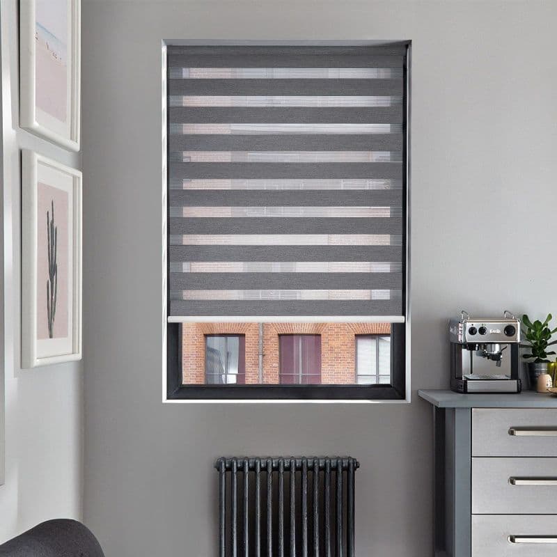 Gray zebra roller blind, lowered with alternating opaque and sheer horizontal bands, filtering light and partially revealing a brick building through the lower pane; kitchen corner with espresso machine, radiator.