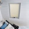 Cream pleated skylight blind, fully lowered with visible dotted ladder tapes, softly filtering daylight through a roof window; set in a white-painted attic bedroom with horizontal wood paneling and pillows.