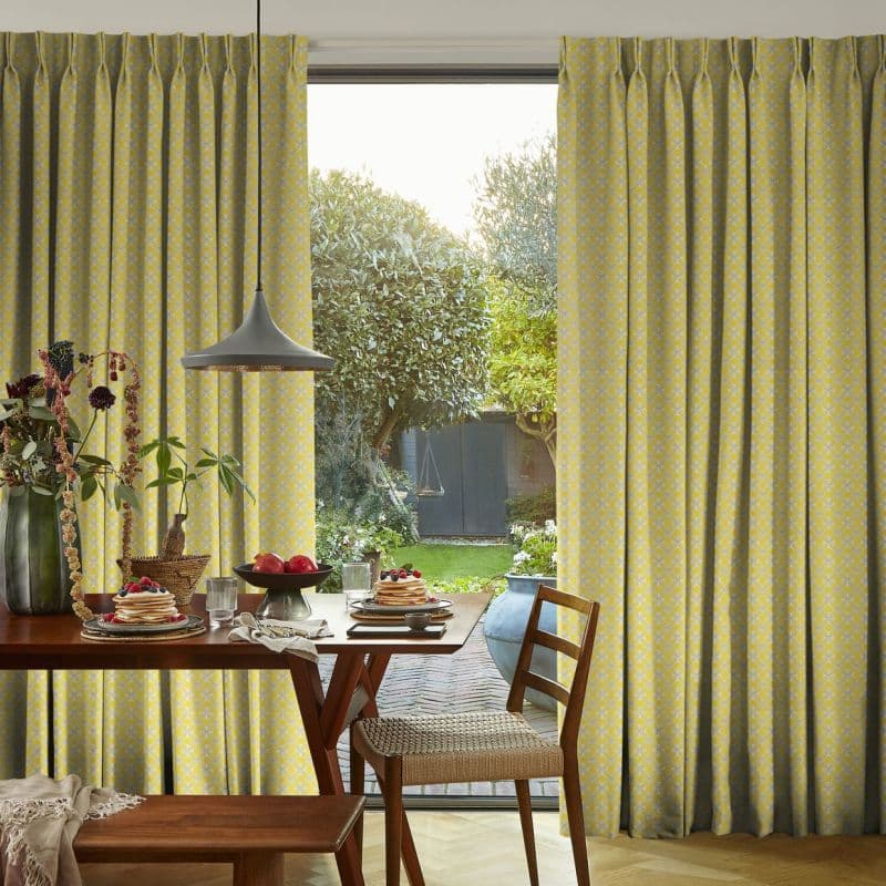 Full-length pleated yellow patterned curtains, parted centrally to reveal a glass door; filtering soft daylight. Dining table set with pancakes and fruit, pendant lamp overhead, view of a green backyard.