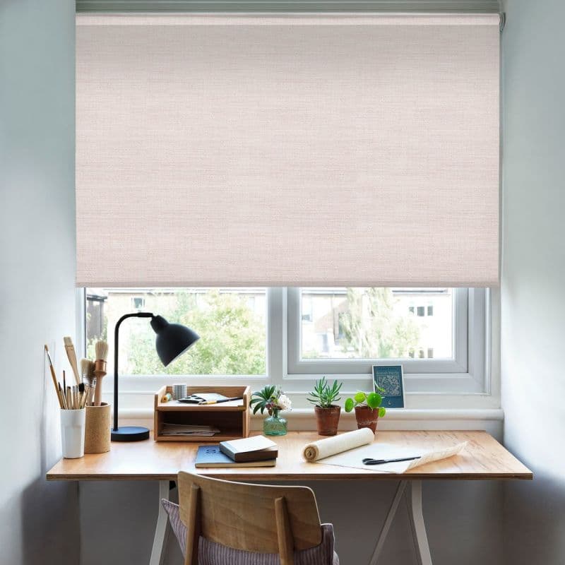 A pale textured roller blind, lowered to cover most of the window, diffusing daylight; a wooden desk beneath holds a lamp, art brushes, notebooks and potted plants in a bright study.