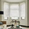 Three white zebra roller blinds lowered across a bay of windows, alternating sheer and opaque bands, filtering soft daylight into a living room with sofa, lamp and coffee table.