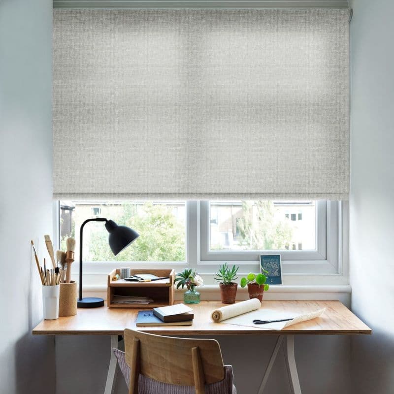 Light-grey textured roller blind lowered almost to the sill, its woven fabric filtering soft daylight; a home workspace sits beneath — wooden desk with lamp, brushes, notebooks and potted succulents.