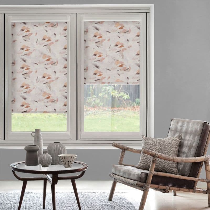 Patterned roller blinds with peach-and-taupe brushstroke print, covering twin windows and partially lowered, filtering daylight; grey-walled living room with rattan armchair with cushion, round coffee table, lawn visible outside.