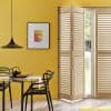 Wooden plantation shutters folded open to the right, bi-fold panels with angled louvers letting daylight through. In a yellow-walled dining area with black pendant lamp, table, chairs, and garden view.