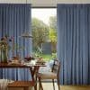 Blue floor-length pleated curtains, hung and parted centrally to reveal sunlight; framing a wooden dining table laid for breakfast in a softly lit room that opens onto a green backyard.