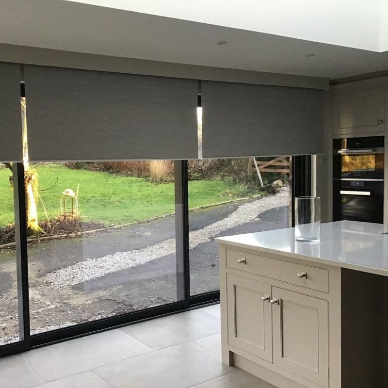 Grey roller blinds lowered across wide sliding glass doors, with narrow vertical gaps filtering light; modern kitchen island with glass cylinder, tiled floor, view of driveway and green lawn outside.