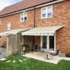 A pair of striped cream-and-green retractable awnings extended over ground-floor French doors and a patio, shading a small outdoor seating area with table and chairs in a sunny brick-backed garden.