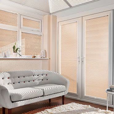 Beige pleated blinds (fitted inside frames) covering bay windows and French doors, closed and filtering sunlight; living room with white tufted sofa, wooden floor, shag rug, daytime light.
