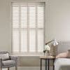 White full-height plantation shutters covering a tall window, louvers tilted to filter soft daylight; set in a neutral-toned bedroom with upholstered bed, bedside table lamp, vase of flowers and armchair.