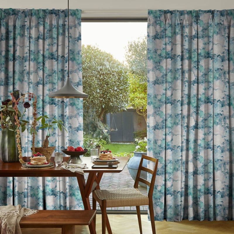Full-length pleated curtains with blue-green watercolor pattern, parted to frame a sliding door; allowing morning light into a dining nook with wooden table, pendant lamp, breakfast plates, and garden view.