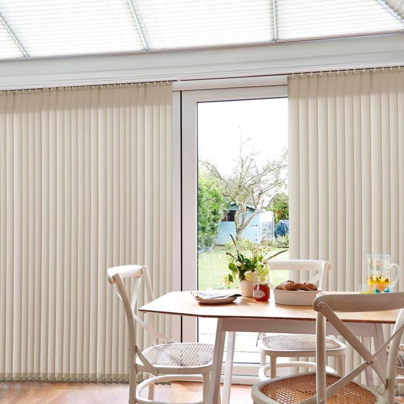 Beige vertical blinds covering patio door, mostly closed with a central opening revealing a garden; they hang straight, filtering daylight into a bright dining area with wooden table and chairs.