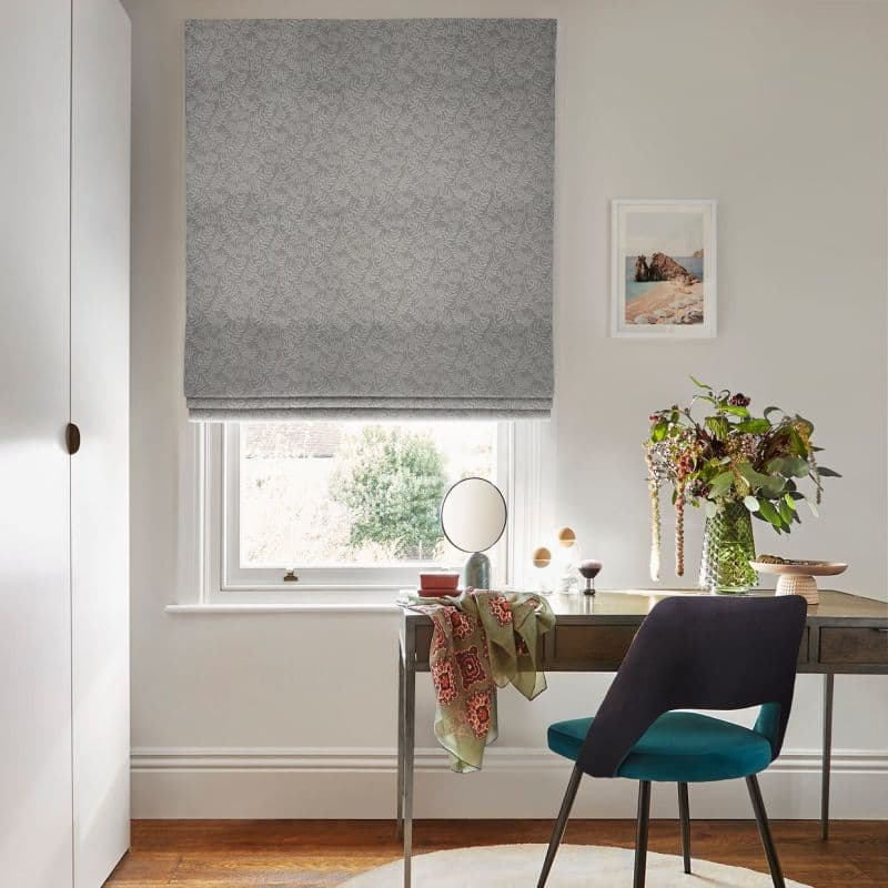 A grey patterned Roman blind lowered over a sash window, filtering daylight; a desk beneath holds a mirror, scarf, vase of flowers and teal-upholstered chair in a lit home office.
